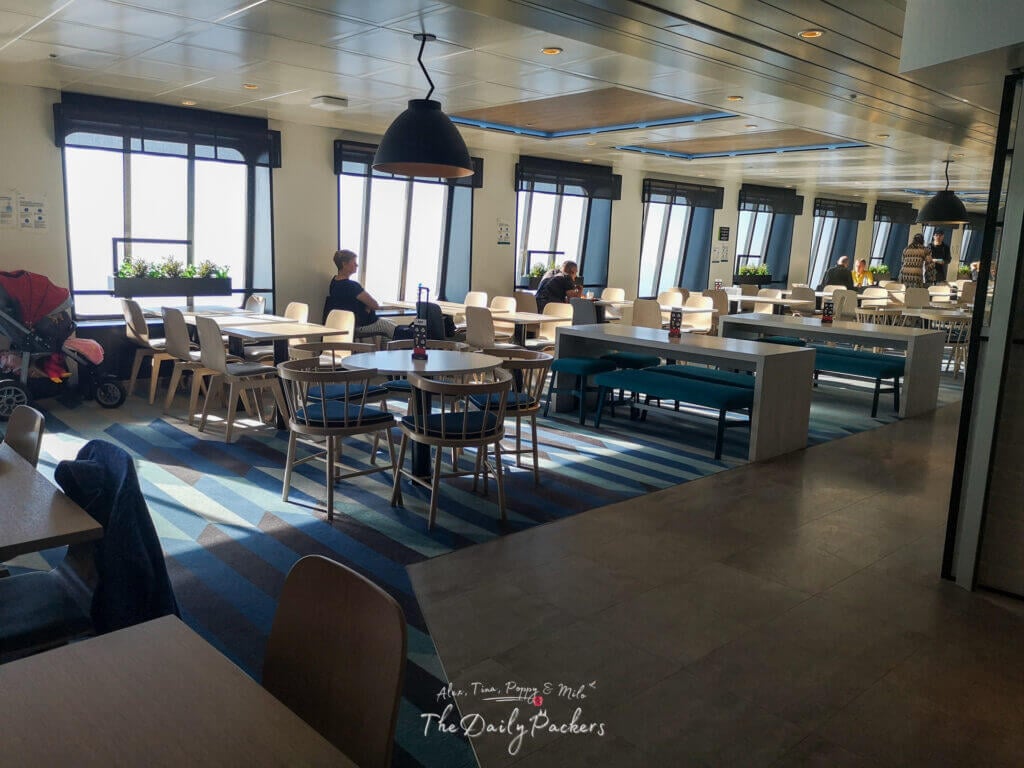 Spacious indoor ferry lounge with large windows, passengers seated at tables, and a family stroller nearby