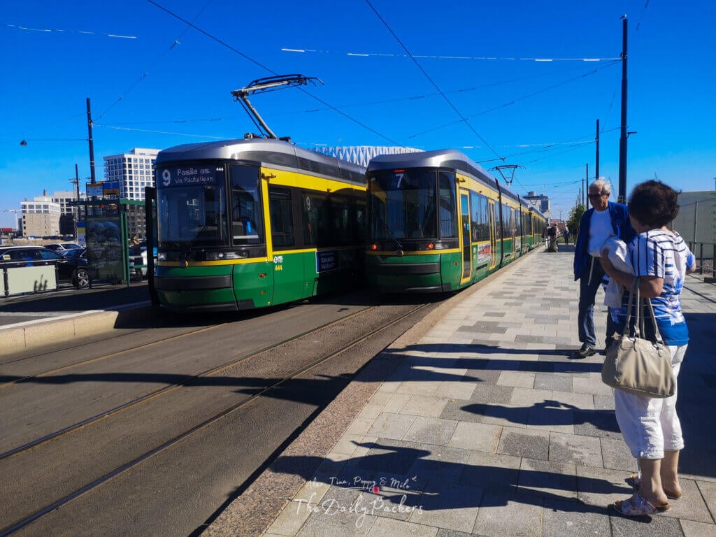 Green and yellow trams at a station in Helsinki with people waiting under a clear blue sky