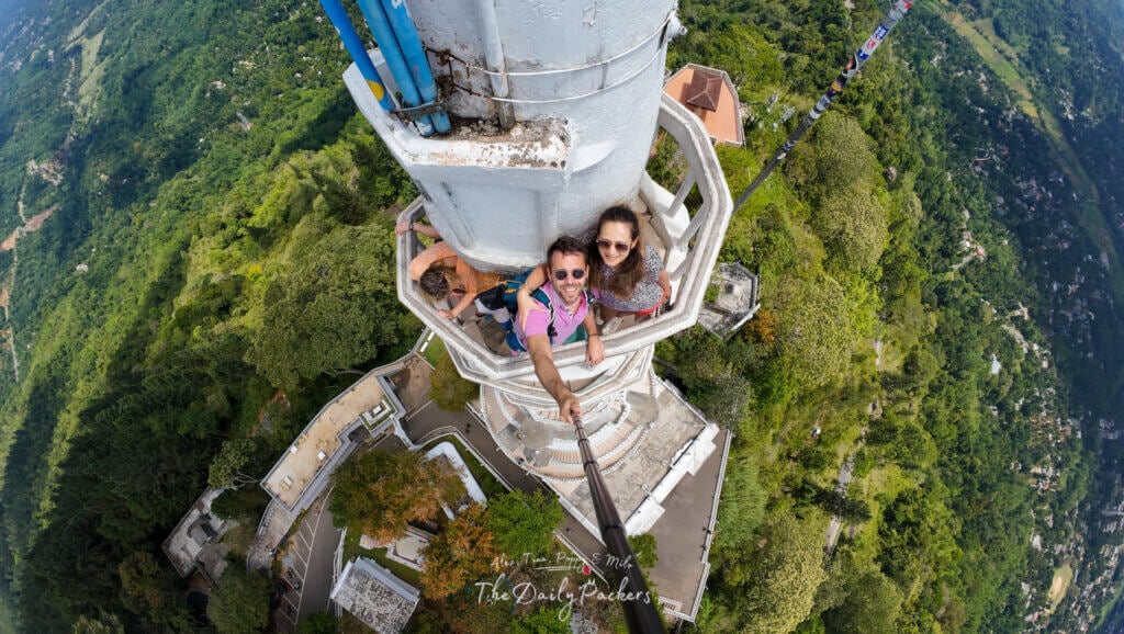 Couple prenant un selfie depuis l'étroit balcon de la tour d'Ambuluwawa au Sri Lanka, avec une vue spectaculaire sur les collines verdoyantes et les routes sinueuses en contrebas.