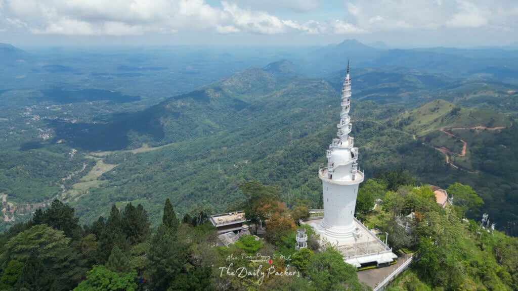 Vue aérienne de la tour d'Ambuluwawa s'élevant au-dessus de collines et de vallées verdoyantes au Sri Lanka, avec des montagnes lointaines sous un ciel partiellement nuageux.