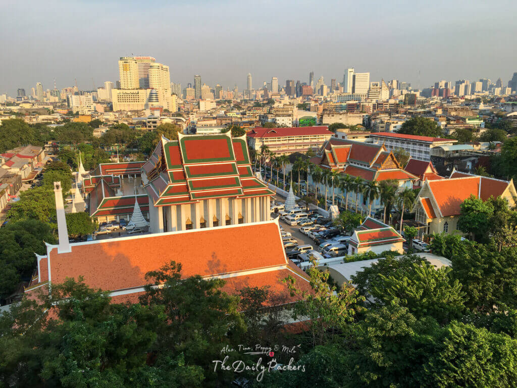 View over Bangkok city from Golden Mount, showing temple rooftops with orange tiles in the foreground and modern high-rise buildings in the distance under a hazy sky.