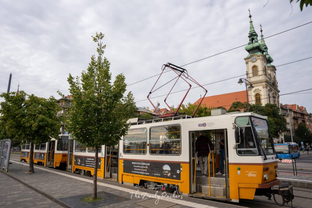 Yellow tram at a city station with church and cityscape in the background in Budapest