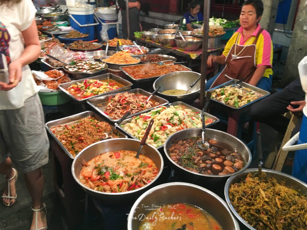Colorful array of Thai street food dishes laid out in metal trays at a busy local market in Chiang Mai.