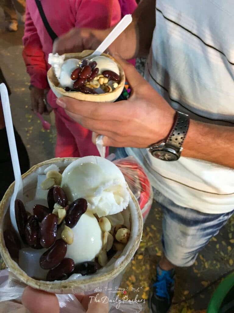 Close-up of Thai coconut ice cream served in a coconut shell with red beans and peanuts, held at a lively night market.