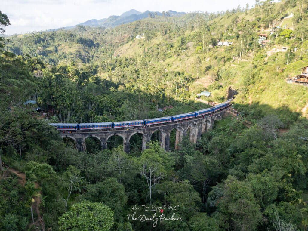 Side aerial shot of the Nine Arches Bridge with a colorful train, rolling through the green hills near Ella.