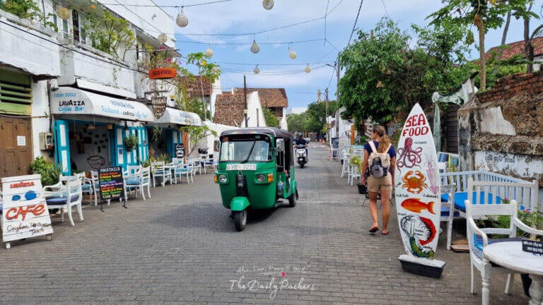 Rue étroite de Galle Fort bordée de cafés blanchis à la chaux et de restaurants de fruits de mer. Un tuk-tuk vert passe devant des sièges en plein air et une femme marche devant.