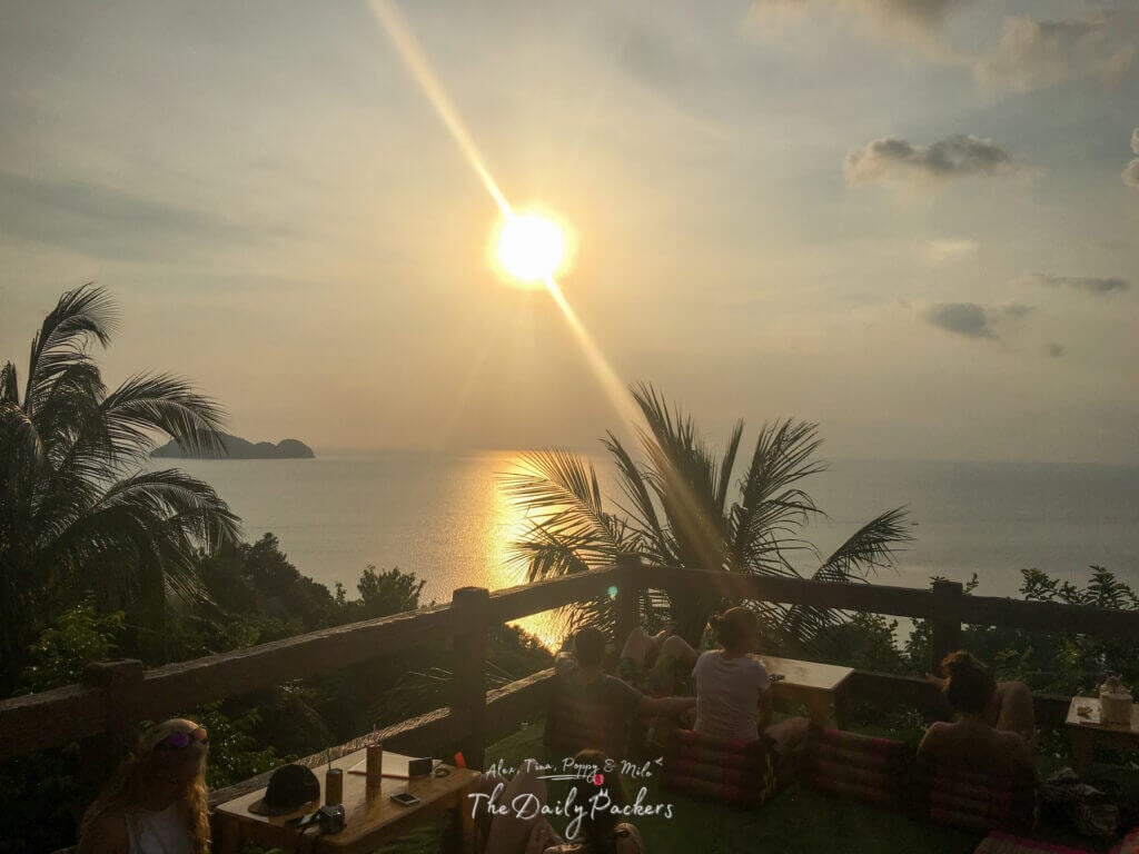 Travelers lounging on floor cushions at Amsterdam Bar on Koh Pha Ngan, watching a golden sunset over the ocean with silhouetted palm trees and calm waters.