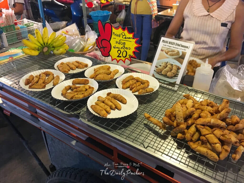 Fried banana rolls displayed on plates at a vibrant Koh Pha Ngan night market stall, with handwritten signs and a vendor preparing more treats in the background.