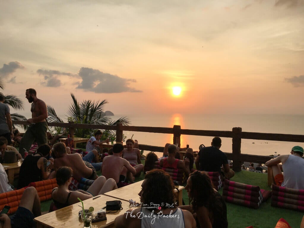 Groups of travelers relaxing at Amsterdam Bar on Koh Pha Ngan, enjoying drinks on outdoor cushions while watching the sun set over the ocean.