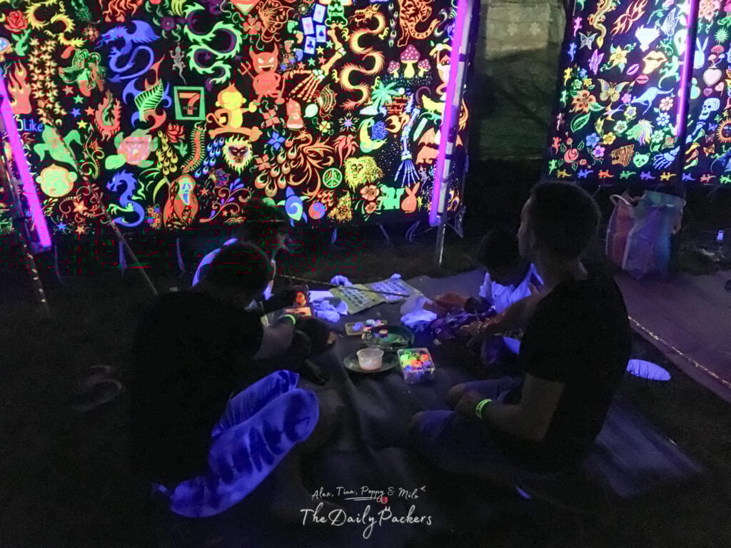People sitting on mats painting under blacklight at the Jungle Experience on Koh Pha Ngan, with neon psychedelic art glowing on surrounding walls.