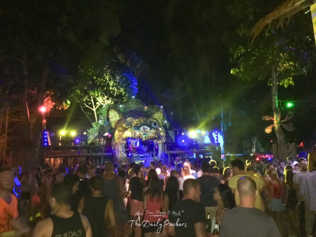 Crowd of people dancing at the Jungle Experience Party on Koh Pha Ngan at night, with colorful lights and a decorated stage surrounded by trees.