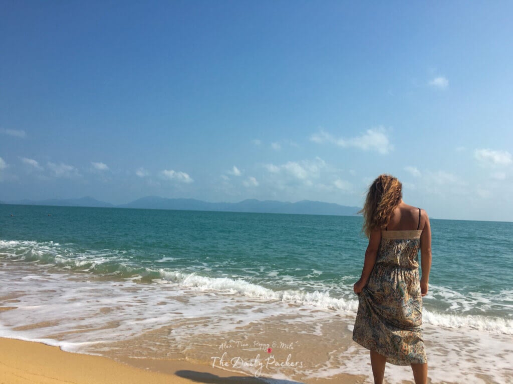 Woman in a summer dress standing at the edge of the water looking at the sea and distant hills.