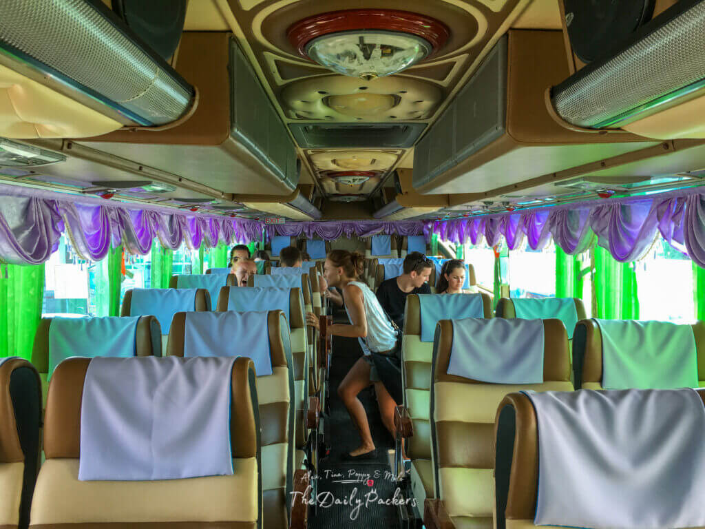 Interior of a colorful Thai bus with green curtains and travelers settling into wide seats.