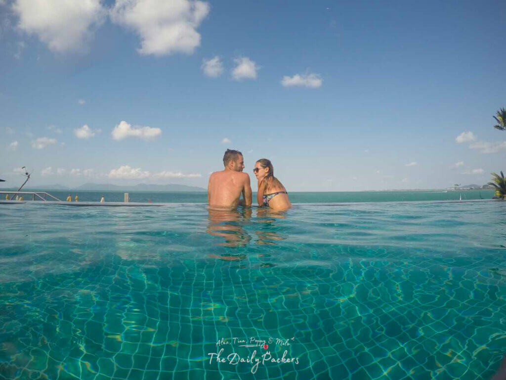 Couple relaxing in an infinity pool with turquoise water overlooking the sea and distant mountains under a blue sky with scattered clouds.