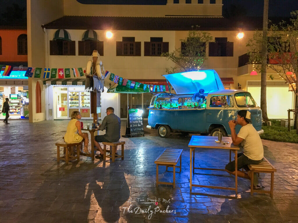Open-air bar in a courtyard at night with customers seated on wooden benches and a converted van serving drinks
