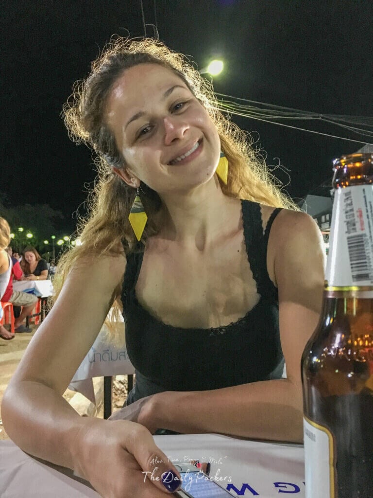 Smiling woman with long earrings sitting at a night market table in Krabi, Thailand, holding a beer with other diners in the background.
