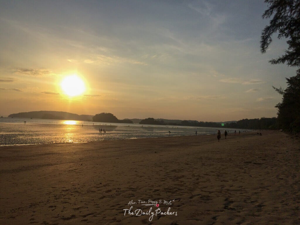 Daytime view of Ao Nang Beach in Krabi, Thailand with gentle waves, a few beachgoers, and tree branches framing limestone cliffs.