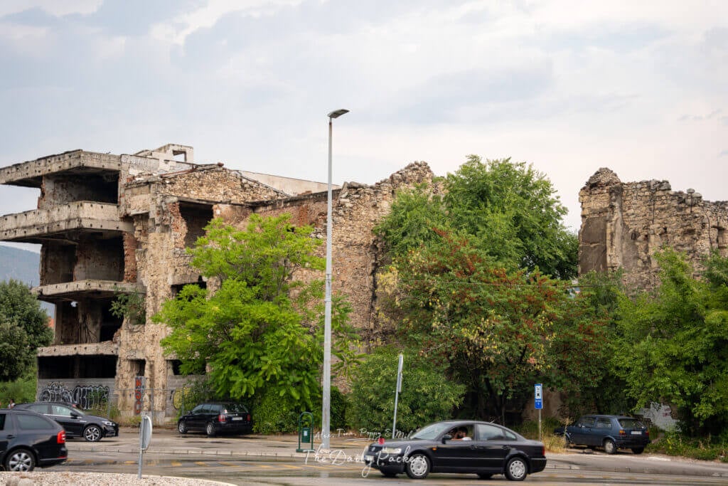 Vestiges d'un bâtiment endommagé par la guerre à Mostar, avec des briques apparentes et des barres d'armature envahies par la verdure.