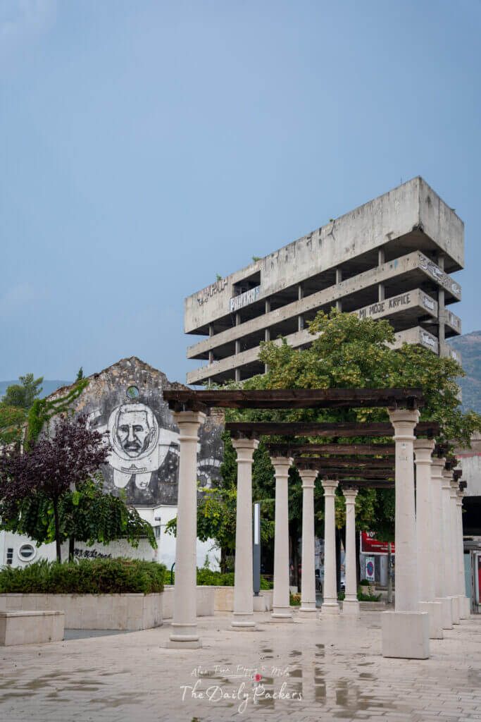 Grandes colonnes blanches du parc de la Place d'Espagne à Mostar, avec en toile de fond des bâtiments endommagés par la guerre et des œuvres d'art de rue.