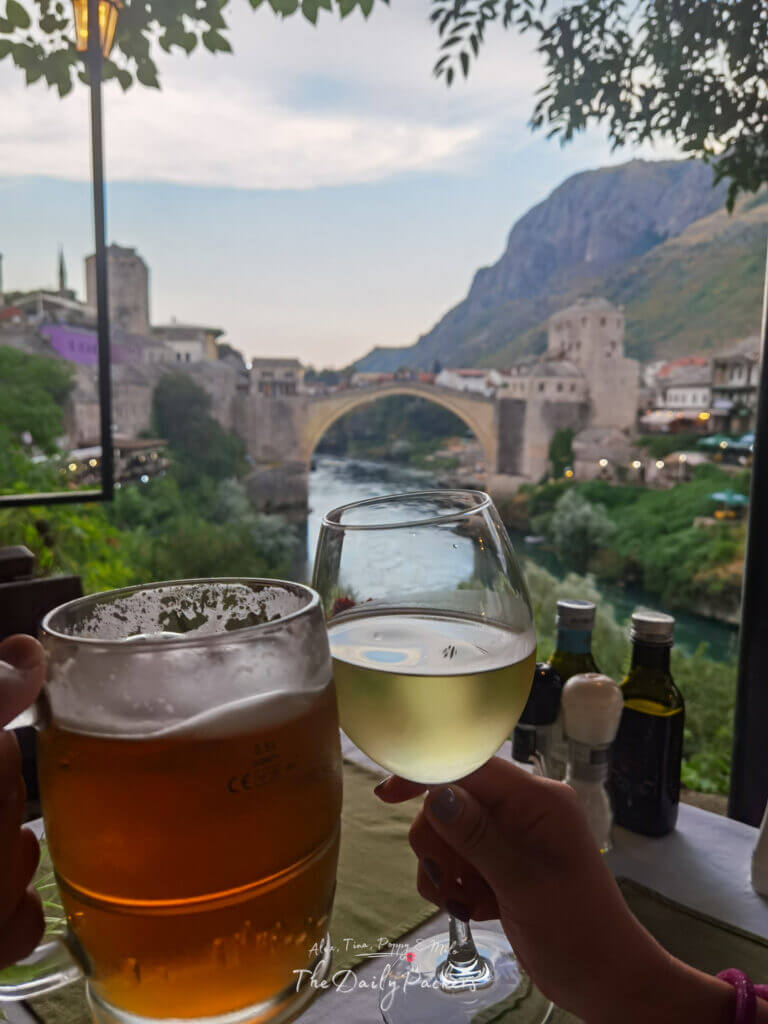 Deux personnes trinquent avec un verre de vin et une bière dans un restaurant avec vue sur le pont Stari Most au coucher du soleil à Mostar, Bosnie-Herzégovine.