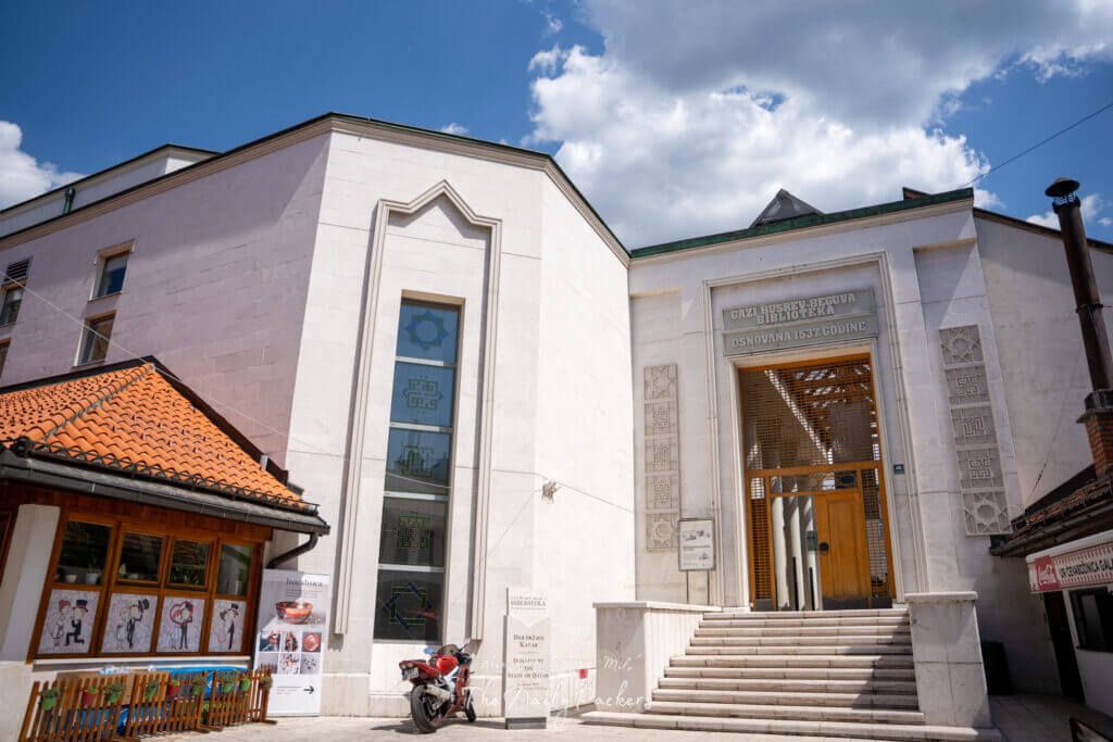Façade blanche moderne de la bibliothèque Gazi Husrev-beg à Sarajevo, avec des fenêtres décoratives et de grandes marches d'entrée sous un ciel bleu.