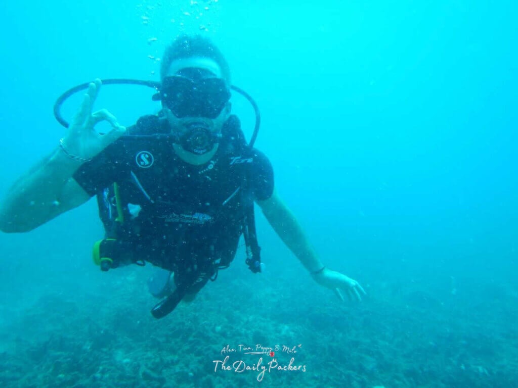 Man in scuba diving gear underwater making an OK hand signal surrounded by blue water and coral on the ocean floor.
