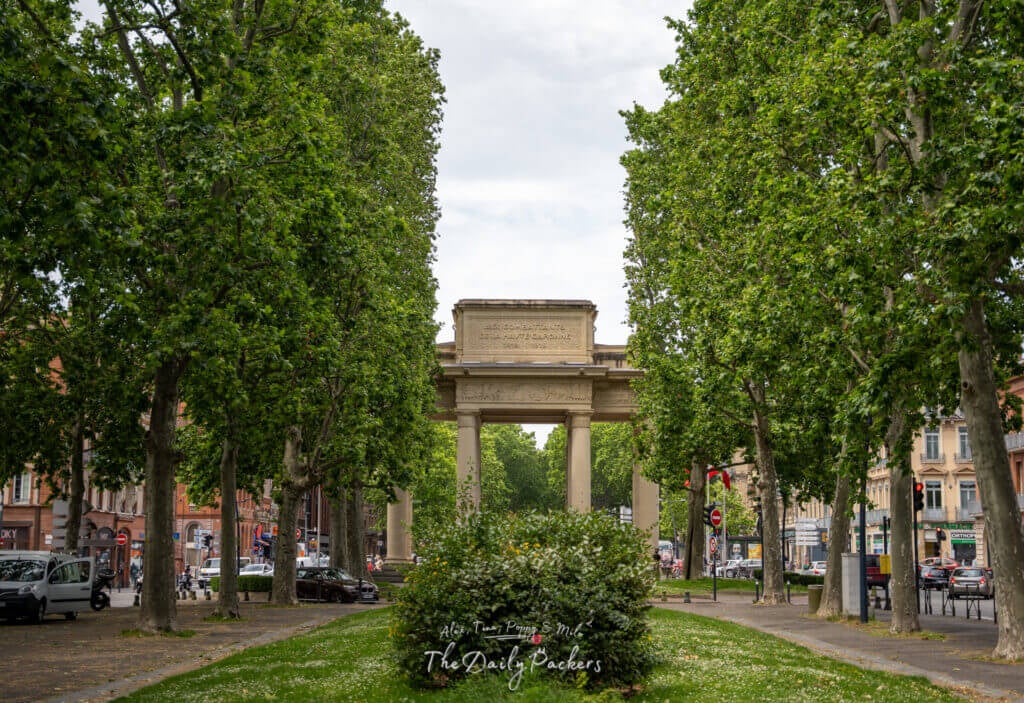 Triumphal arch surrounded by lush trees and people walking near the entrance of Toulouse’s