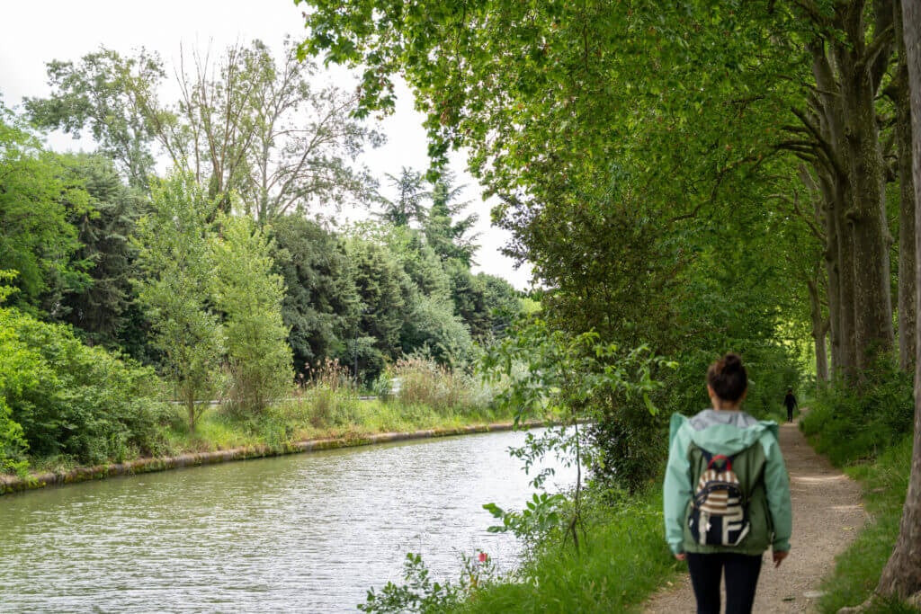Woman walking along the shaded Canal du Midi path bordered by tall trees and calm waters.