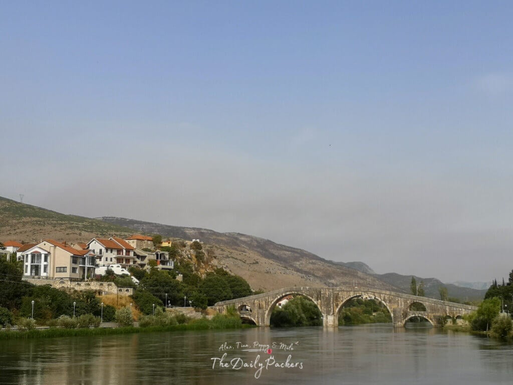 Panoramic view of the Arslanagić Bridge crossing the Trebišnjica River surrounded by hills and houses.