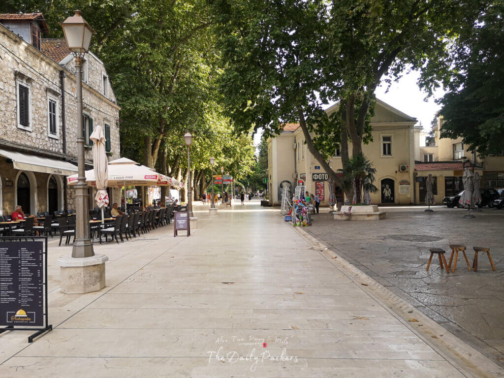 Main square in Trebinje with cafés and restaurants shaded by tall trees, featuring stone buildings and outdoor seating.