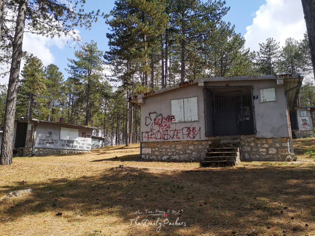 Close-up view of a rundown cabin with graffiti in the forested remains of a refugee camp.
