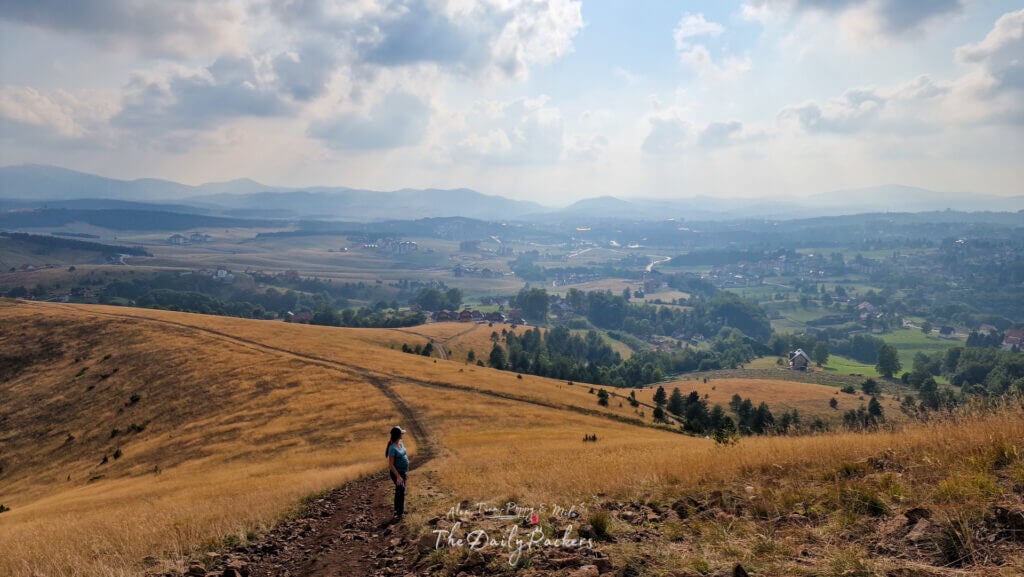 Woman walking along a dirt path through rolling hills with Zlatibor town and misty mountains in the background