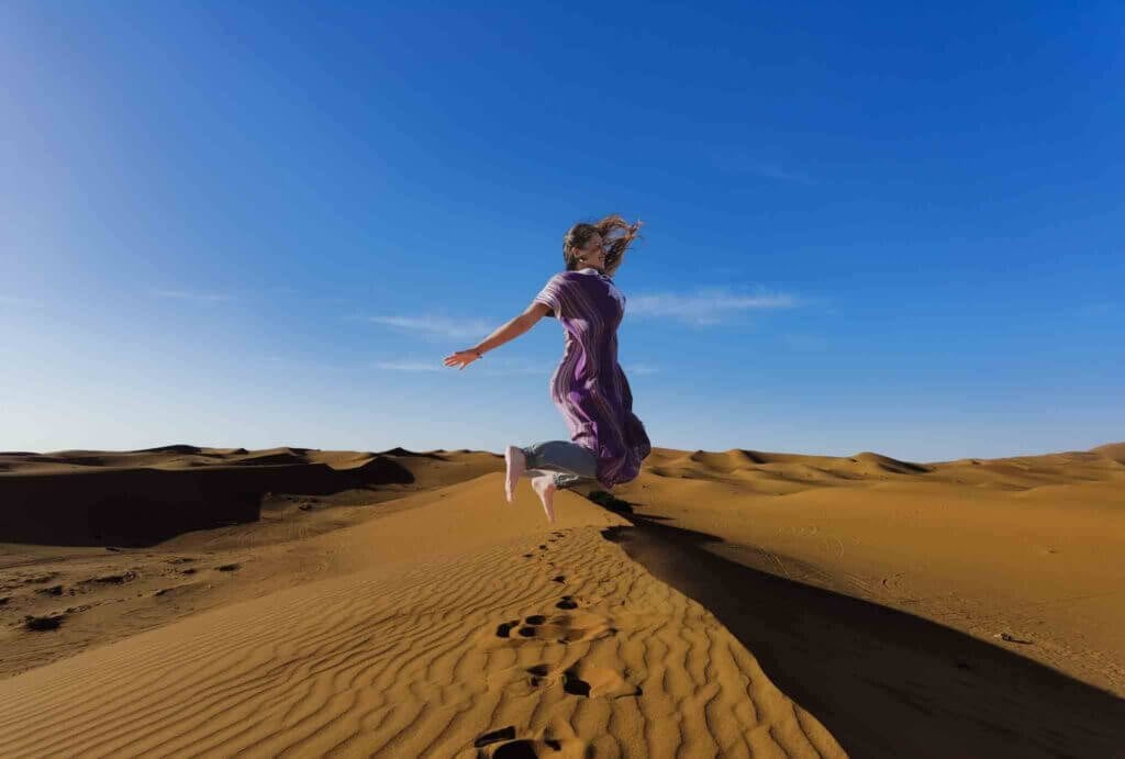 femme sautant pieds nus sur des dunes de sable sous un ciel bleu clair au Maroc