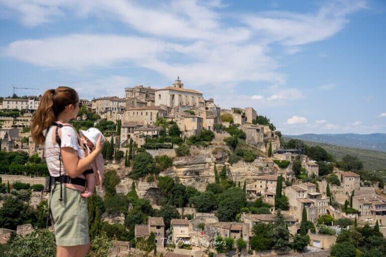 Woman holding baby while looking out at the dramatic stone houses of Gordes cascading down the hillside