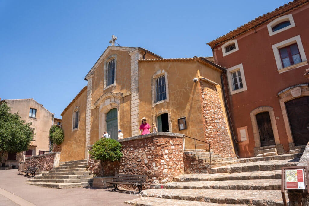Église peinte en ocre avec des marches en pierre et des visiteurs à l'extérieur lors d'une journée ensoleillée à Roussillon.