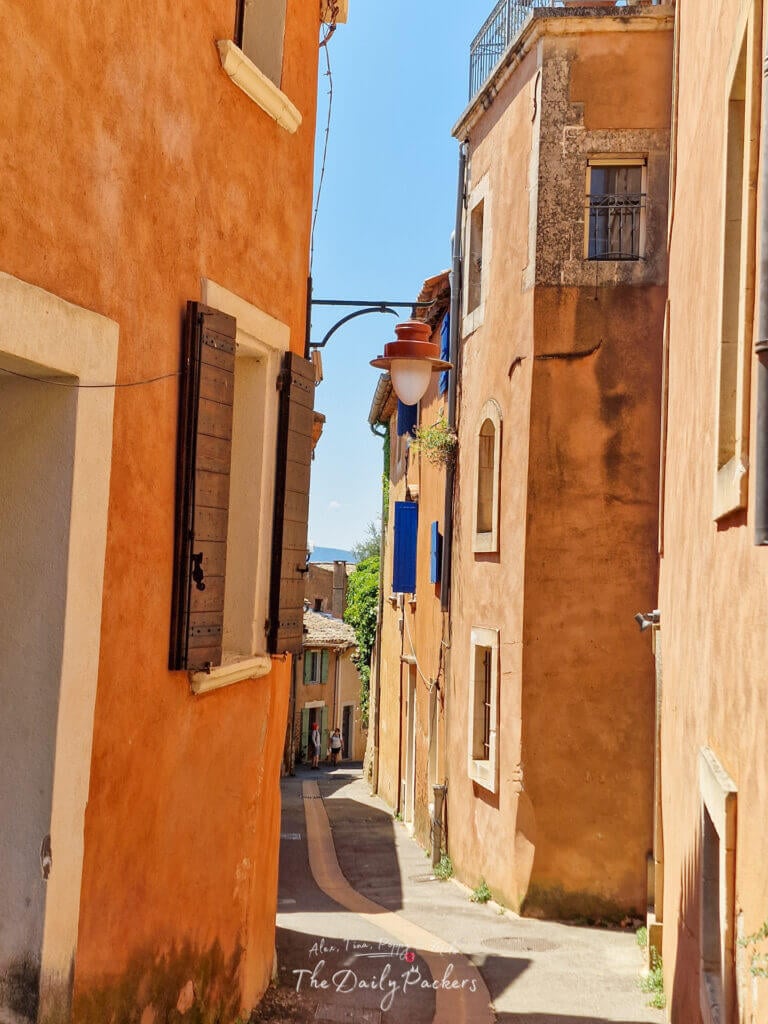 Ruelle du Roussillon encadrée par des bâtiments aux tons chauds, avec des volets rustiques et un lampadaire suspendu au-dessus.