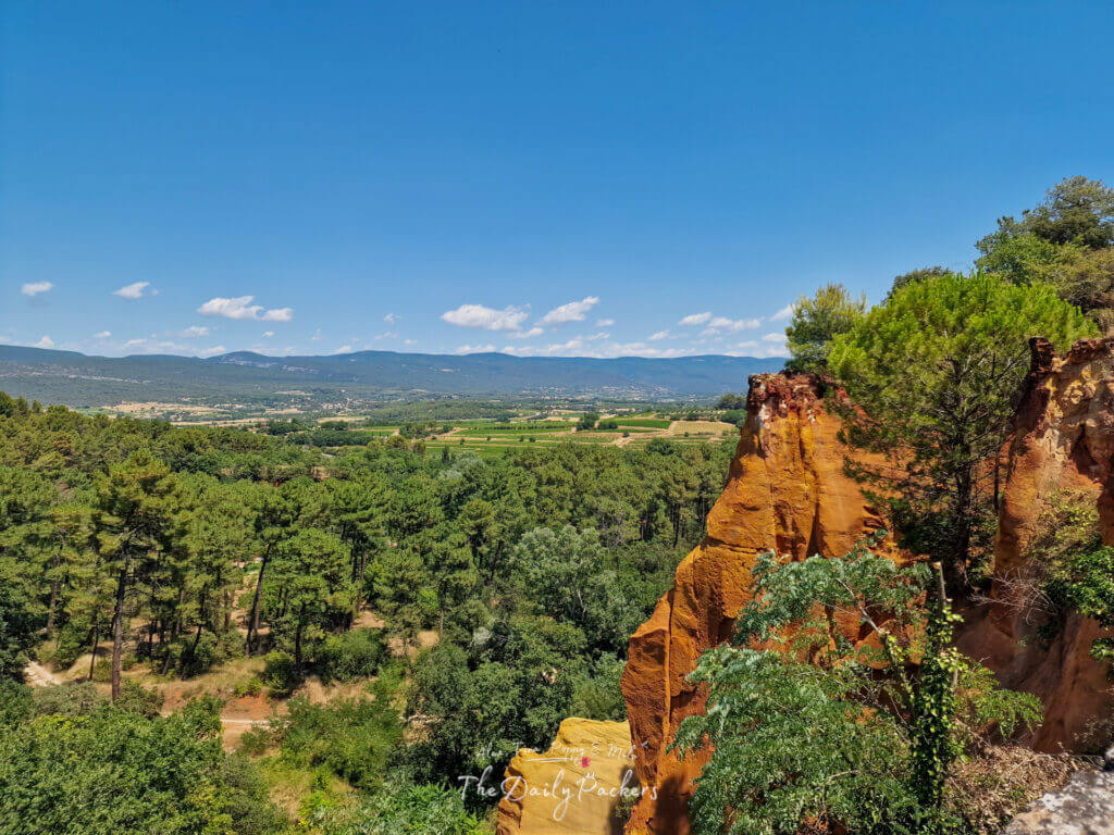 Falaises ocre à l'extérieur de Roussillon avec des pins et des montagnes lointaines sous un ciel clair de Provence.