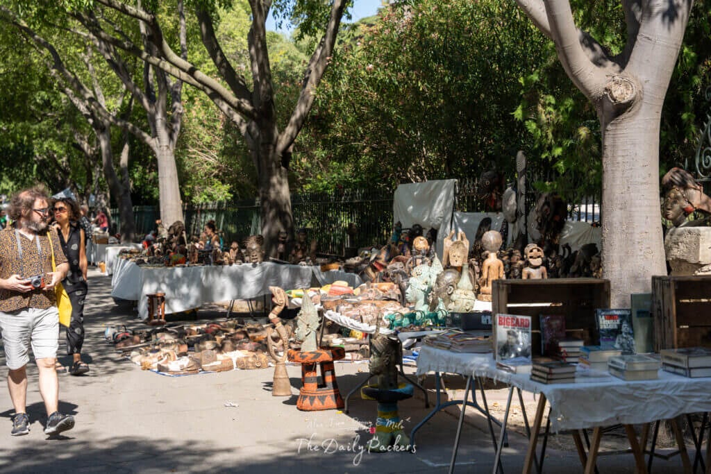 Marché aux puces en plein air à Arles avec des tables remplies d'antiquités, de masques et de sculptures à l'ombre des arbres.