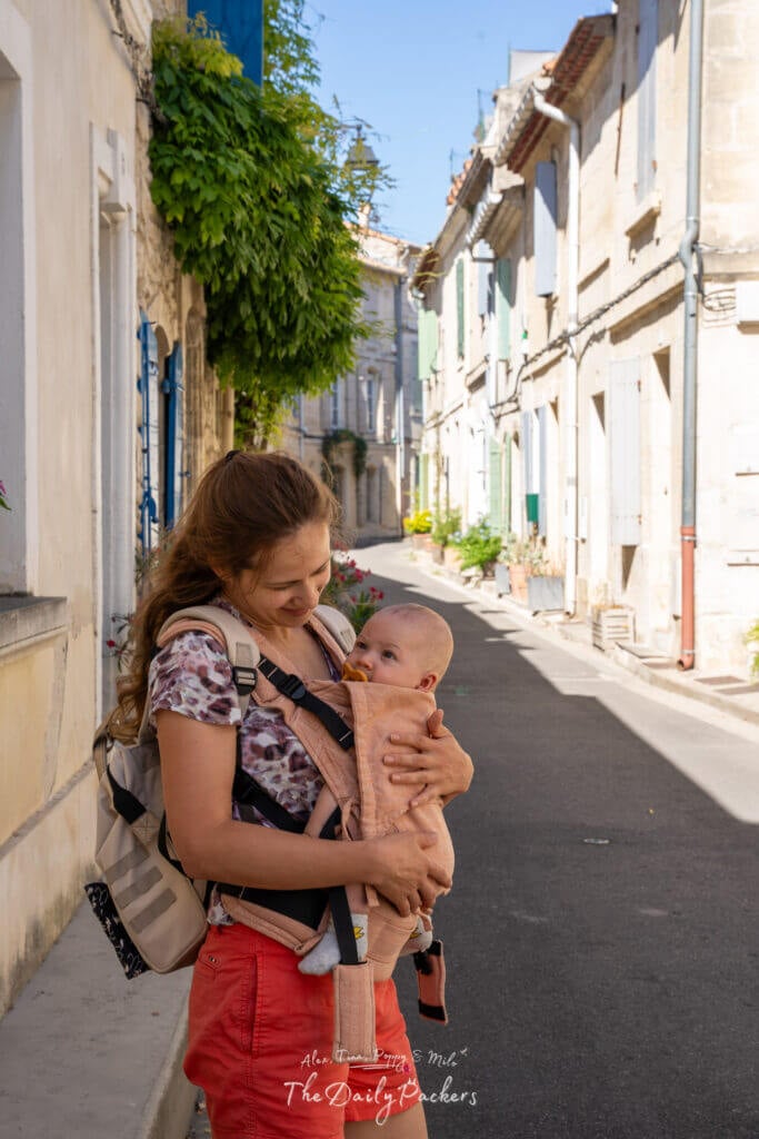 Femme tenant un bébé dans un porte-bébé, souriante dans une rue calme d'Arles aux volets pastels et à la verdure.