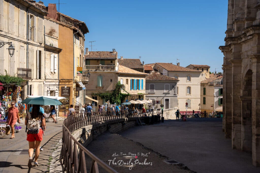 Scène de rue à Arles avec de vieux bâtiments, des terrasses de café et une femme marchant sous un parapluie bleu.