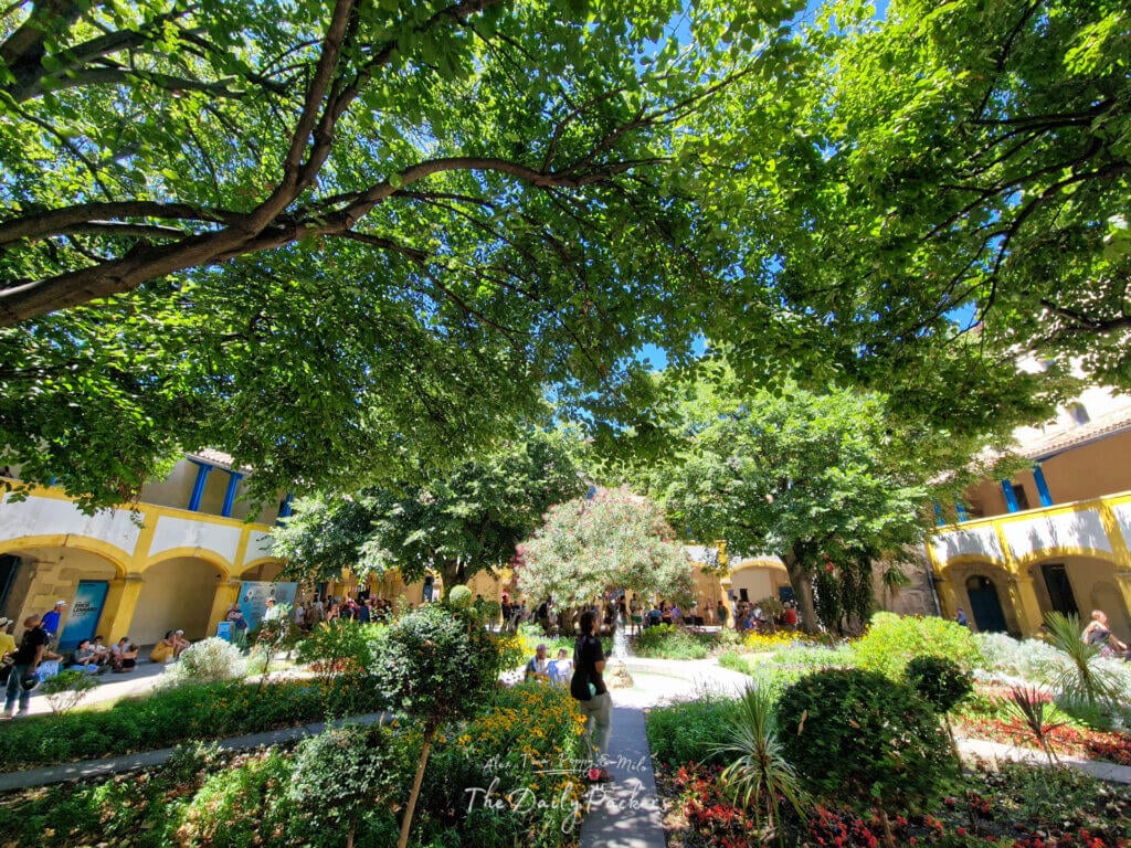 Cour luxuriante de la Maison de Santé d'Arles, entourée de murs aux arches jaunes et de visiteurs qui se promènent.