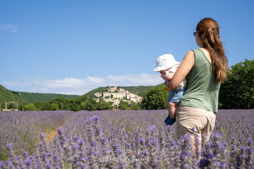 Mère tenant son bébé dans un champ de lavande avec le village de Banon s'élevant au sommet de la colline derrière elle