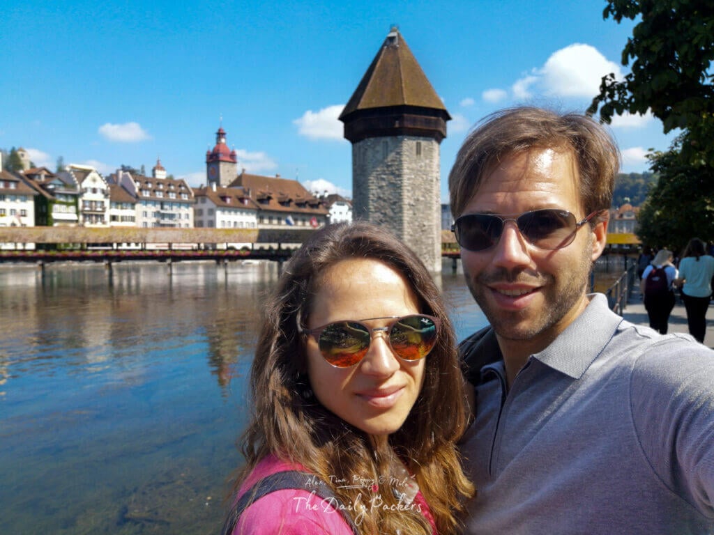 Couple taking a selfie near Chapel Bridge in Lucerne with the water tower and historic buildings behind them.