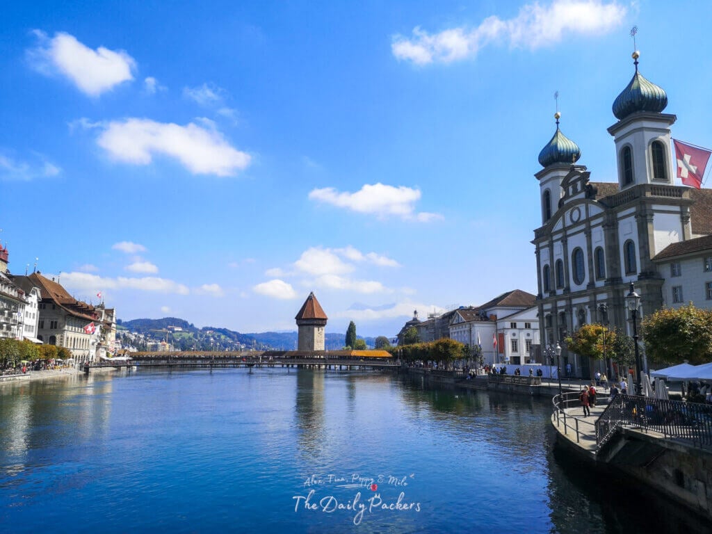 View of the Reuss River in Lucerne with the Jesuit Church and Chapel Bridge in the distance on a bright sunny day.