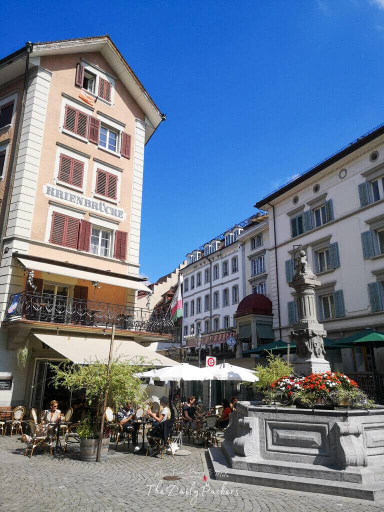 Outdoor café in Lucerne’s city center with a stone fountain decorated with colorful flowers and people dining under umbrellas.