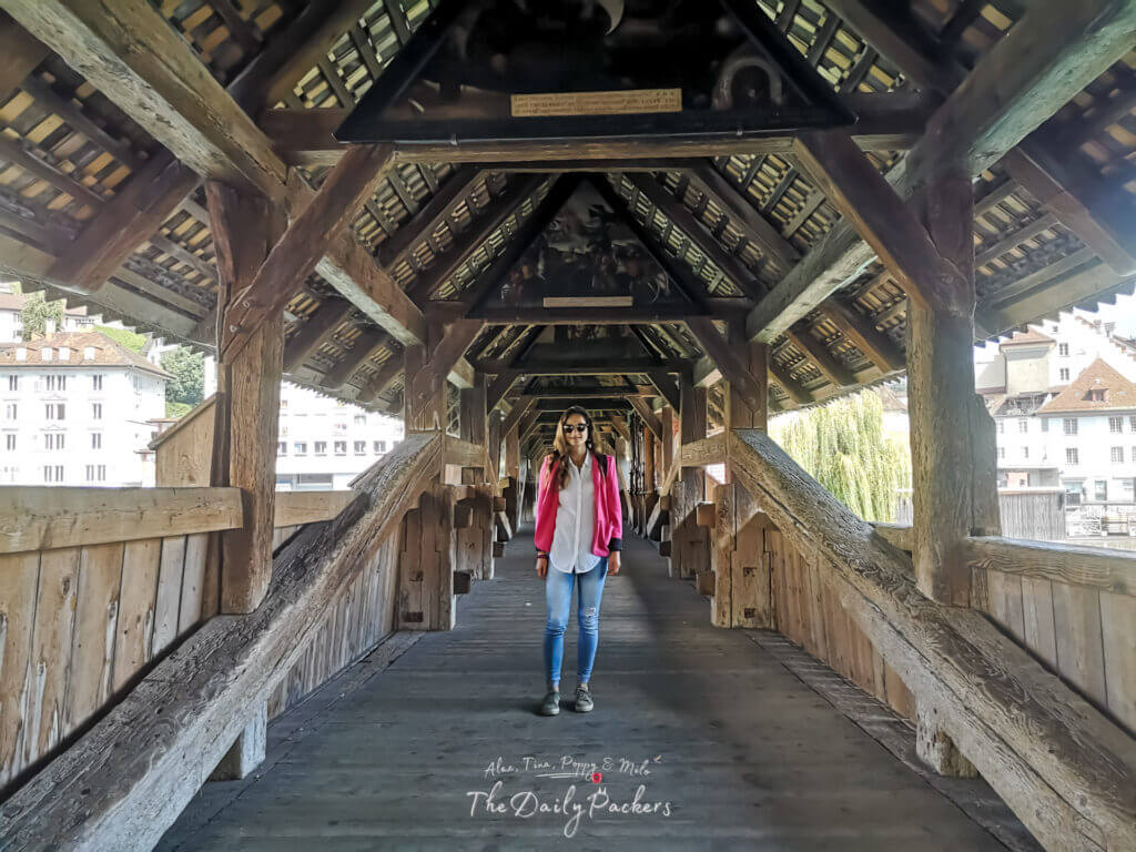 Woman walking inside the covered wooden Spreuer Bridge in Lucerne with its slanted beams and historic paintings overhead.