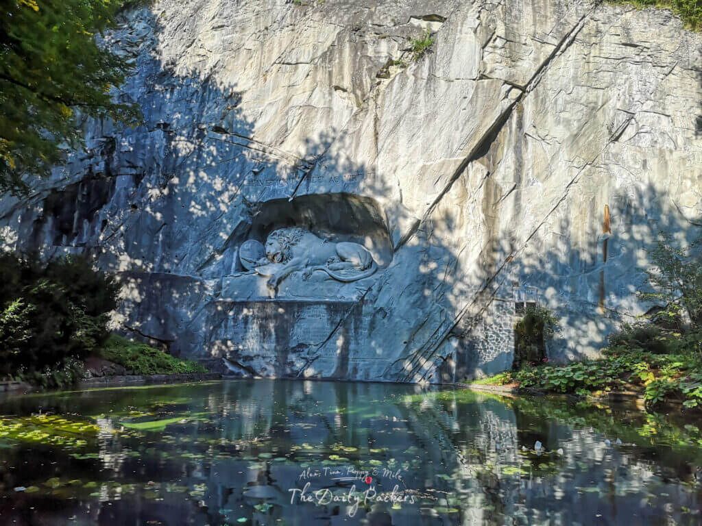 Famous Lion Monument in Lucerne carved into a rock face, reflected in a tranquil pond with scattered leaves.