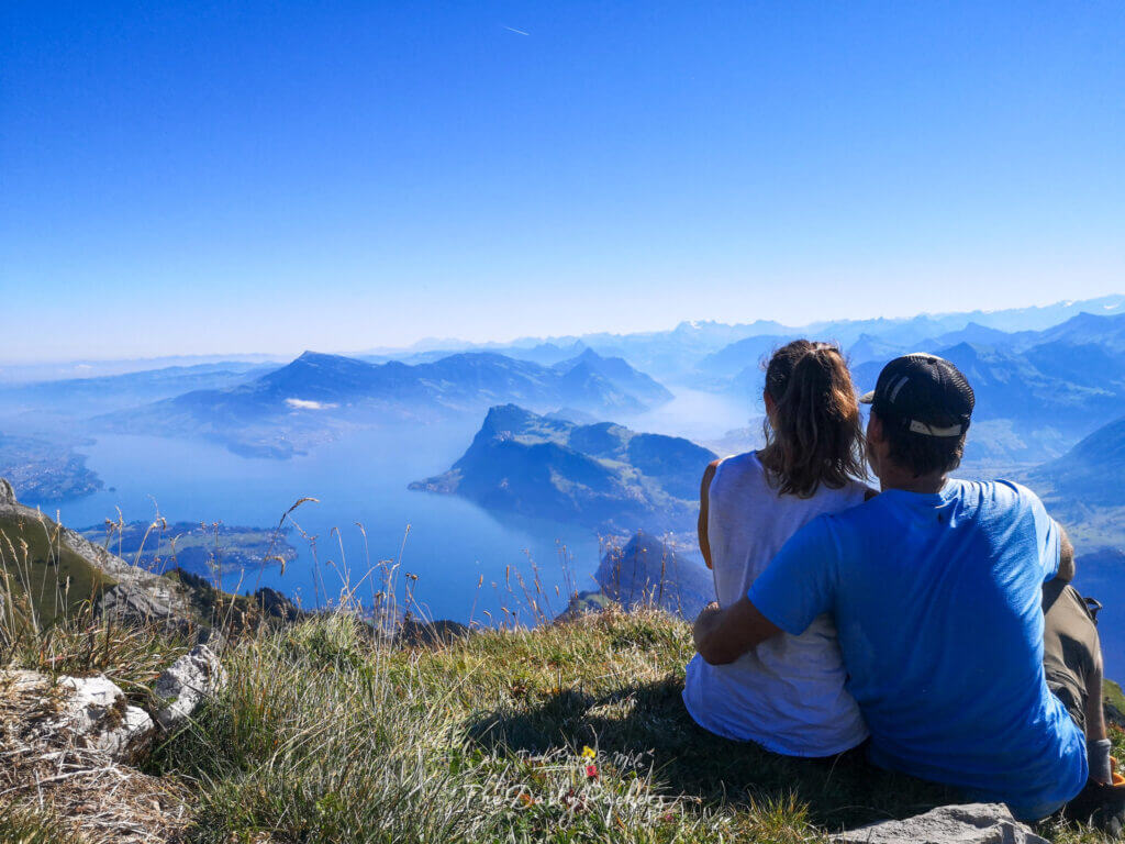 Couple sitting on Mount Pilatus summit, overlooking Lake Lucerne and layered blue Alps under a clear sky.