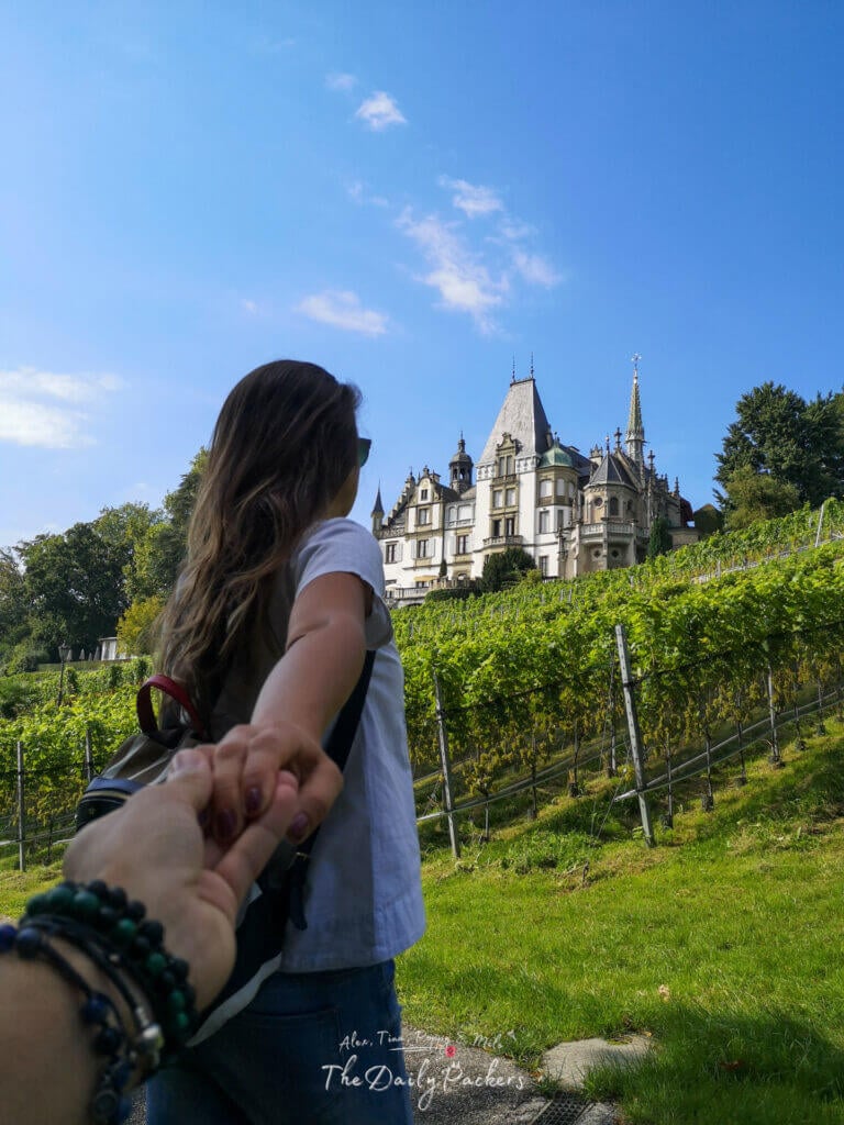 woman holding partner’s hand walking toward Meggenhorn Castle through green vineyards under a bright blue sky