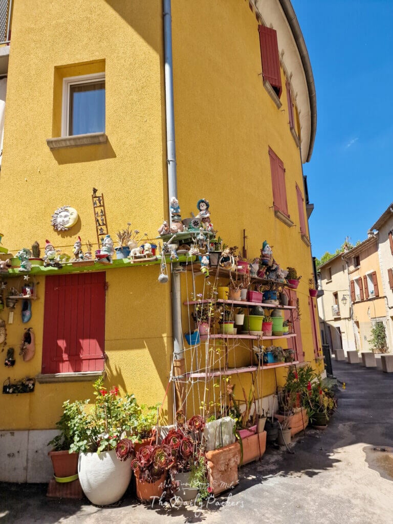 Bright yellow house in Manosque decorated with red shutters, flower pots, and garden figurines.